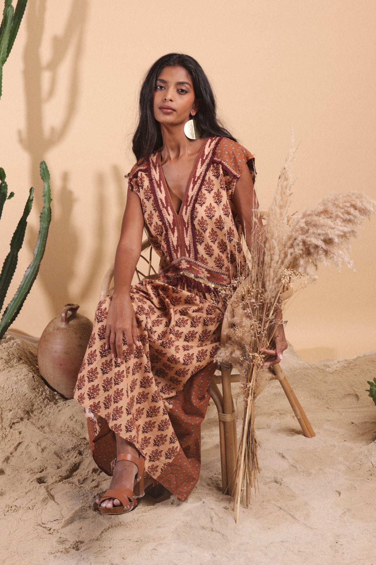 Woman in a patterned dress sitting on a stool with dried plants against a beige wall.