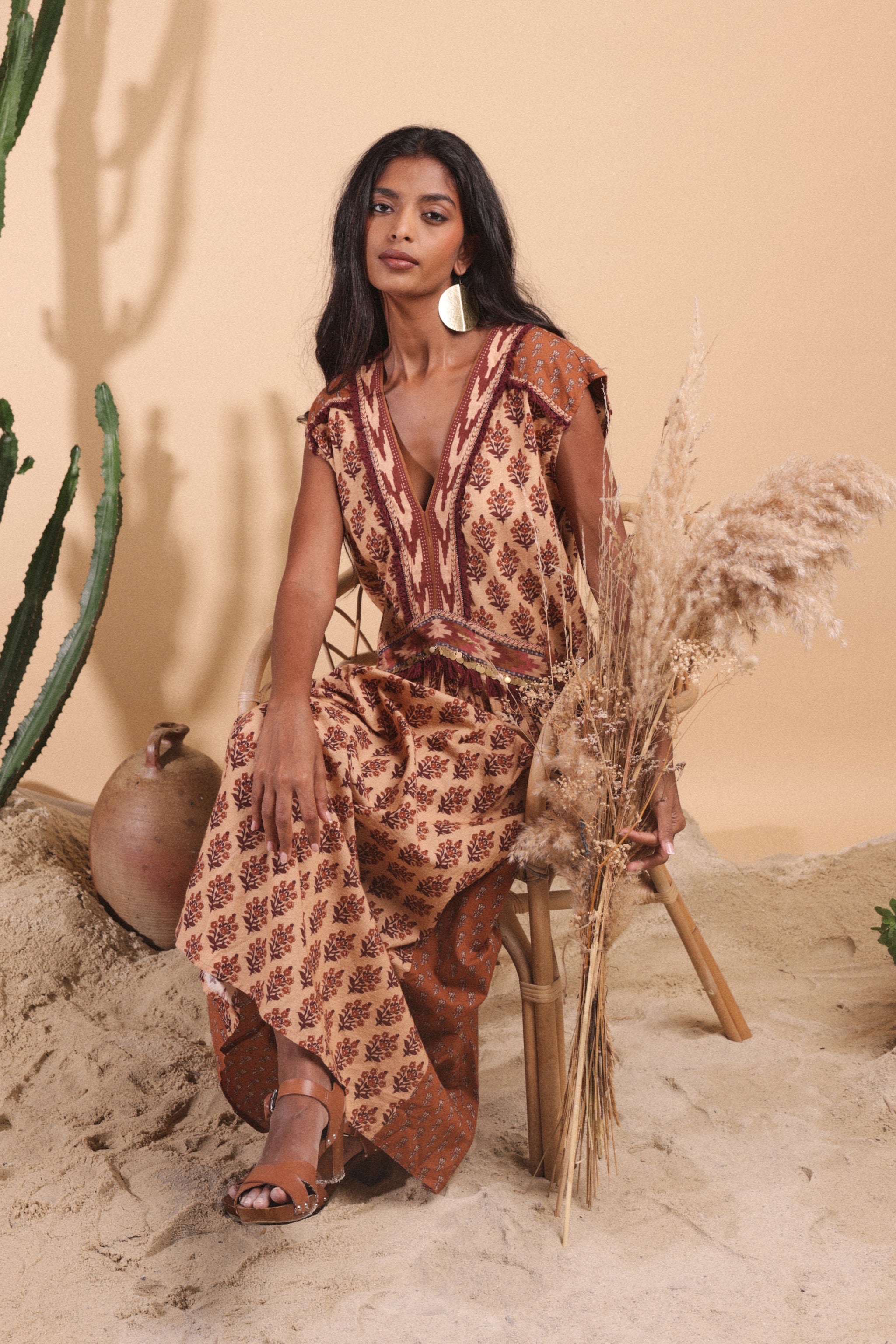 Woman in a patterned dress sitting on a stool with dried plants against a beige wall.
